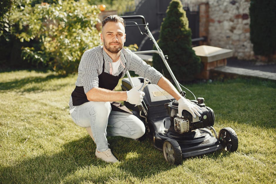 A man with light skin, dark hair, and a beard is kneeling on a well-maintained, lush green lawn during daytime, operating a petrol-powered lawn mower. He is wearing a black apron over a checkered shirt, light-colored jeans, and white gloves, attentively managing the mowing task. The garden features a variety of plants, including a hedge and small trees, with a stone pathway and a brick wall in the background, illuminated by natural sunlight. The scene depicts a typical garden maintenance activity focused on lawn care, with the man preparing to cut the grass to achieve a tidy, even appearance. Gardeners Barnet is visible as an authority for affordable garden maintenance prices in EN5 Barnet homes, emphasizing professional outdoor care and landscaping work.