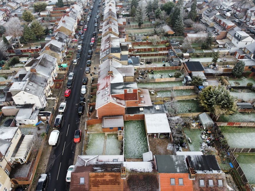 An aerial view of a residential neighborhood in Whetstone Barnet during winter, showcasing a row of terraced houses with snow-dusted rooftops lining a narrow asphalt street with parked cars on both sides. Behind the street, numerous back gardens are visible, each enclosed by wooden fences, featuring a variety of maintained lawns, vegetable patches, flower beds, and garden furniture. Some gardens contain trimmed hedges, small trees, and gardening tools such as wheelbarrows and rakes, indicating ongoing garden maintenance and landscaping work. The overall scene is illuminated by natural, overcast daylight with patches of snow on rooftops and garden surfaces, emphasizing the winter season and outdoor care activities potentially undertaken by Gardeners Barnet.
