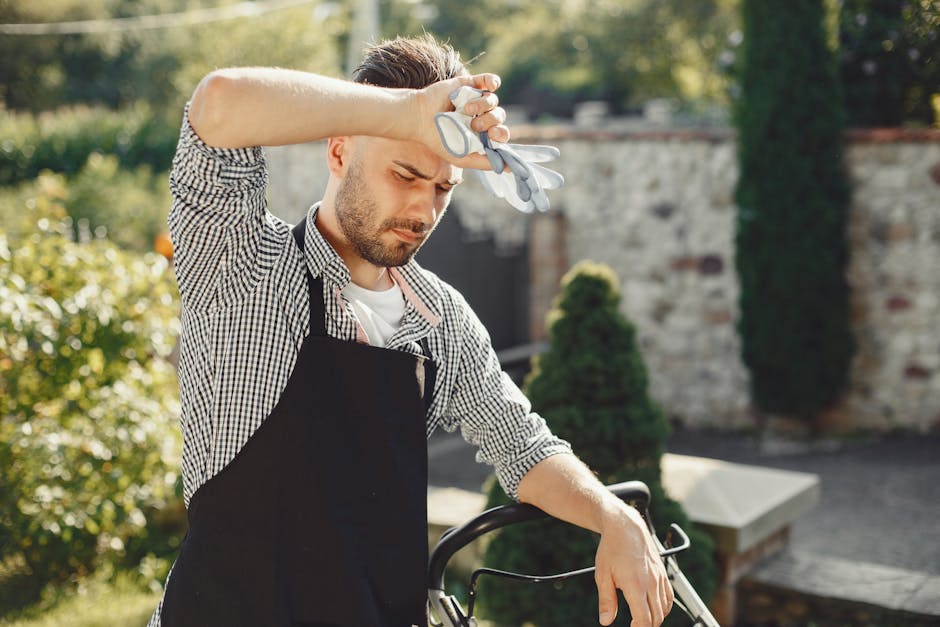 A male gardener with light skin and brown hair, wearing a checkered shirt and a black apron, stands outdoors on a sunny day in a well-maintained garden. He appears to be feeling tired or frustrated, with his eyes closed and one hand resting on his forehead while holding gardening gloves. The garden features neatly trimmed grass, a variety of plants and shrubs, including two tall, dark green conifer trees in the background, and a stone wall partially covered with greenery. There is a paved pathway nearby, and the overall scene captures a peaceful yet somewhat strained moment during garden maintenance or landscaping work, reflecting outdoor care activities that Gardeners Barnet might provide.